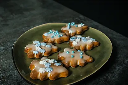 Zimt-Frischkäse-Hörnchen auf einem grünen Teller, dekoriert mit Zuckerguss und bunten Streuseln in Schneeflockenform.