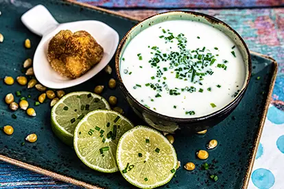 Kalte Buttermilchsuppe mit Limettenscheiben, frischem Schnittlauch und gebackenem Raviolo auf einem dekorativen Teller.