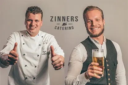 Zwei Männer in professioneller Kleidung lächeln, einer im Kochoutfit mit Daumen hoch, der andere mit einem Bierglas.