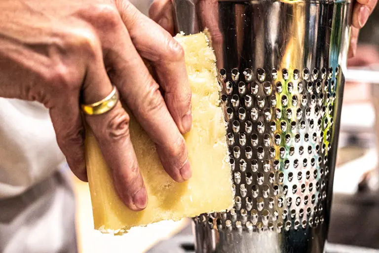 Eine Hand reibt ein Stück Käse über eine Edelstahlreibe im Milchworkshop.