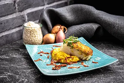 Kabeljau mit Bergkäsekruste auf Chorizo-Safranrisotto, umgeben von Zwiebeln und Risottoreis.