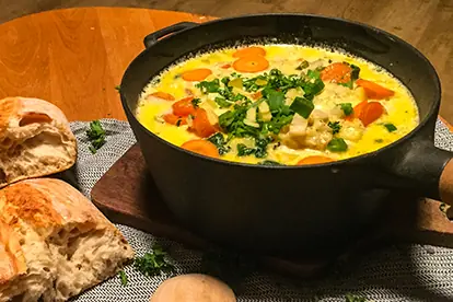 Käsesuppe in einem schwarzen Topf, garniert mit frischen Kräutern und Karotten, neben frischem Brot auf einem Holzbrett.