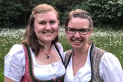 Zwei Frauen in traditioneller bayerischer Tracht lächeln vor einer blühenden Wiese.