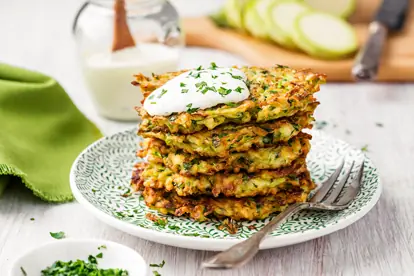 Zucchinipuffer auf einem Teller, garniert mit Kräuterdip und frischen Kräutern, serviert mit einem grünen Tuch.