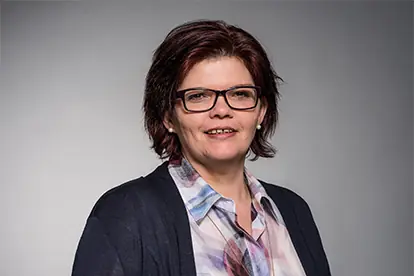 Eine lächelnde Frau mit Brille und blauer Jacke steht vor einem neutralen Hintergrund.