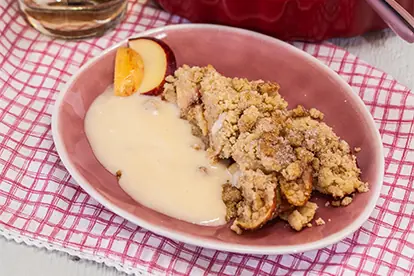 Apfel-Crumble mit Vanillesoße auf einem rosa Teller, serviert mit Apfelscheiben und einem karierten Tischläufer.