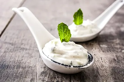 Joghurt-Minze-Dip in weißen Löffeln, garniert mit frischen Minzblättern auf einem rustikalen Holztisch.