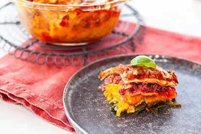 Lasagne mit buntem Gemüse auf einem Teller, serviert neben einer Auflaufform auf einem roten Stoffuntergrund.