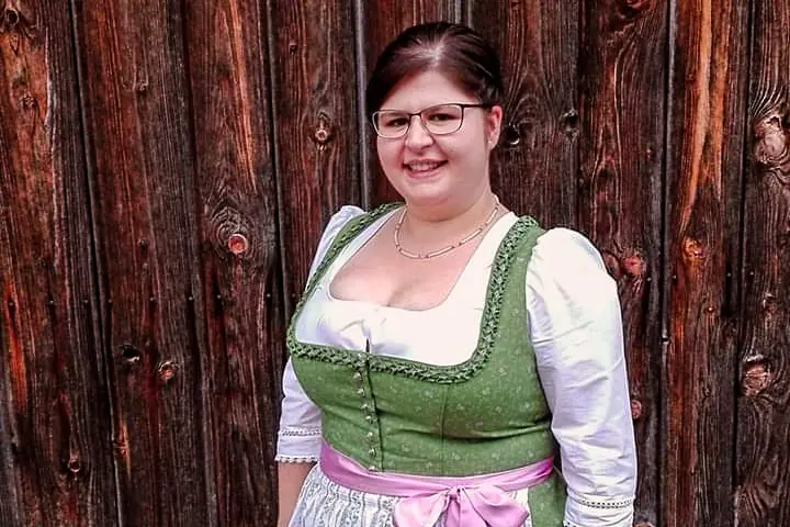 Frau in traditioneller Tracht mit grünem Mieder und rosa Schürze vor einer rustikalen Holzwand.