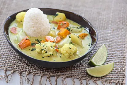 Bunte Gemüsesuppe mit Reis und Limetten, serviert in einer schwarzen Schüssel auf einem rustikalen Untergrund.