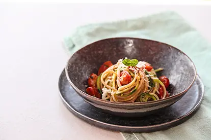 Ein Teller Dinkelsalat mit Spaghetti, Kirschtomaten und frischen Kräutern auf einem grauen Unterteller.