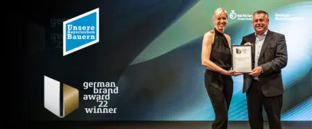 Zwei Personen überreichen einen Preis beim German Brand Award 2022, mit dem Logo und Hintergrundgrafik im Fokus.