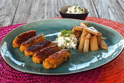 Zucchini-Schiffchen mit Quinoa, Sauerrahm-Dip und gebratenen Kartoffelstücken auf einem bunten Teller serviert.