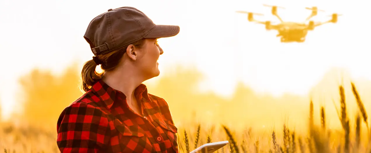 Eine Landwirtin steuert einen Drohne über ein Feld, während die Sonne im Hintergrund untergeht.
