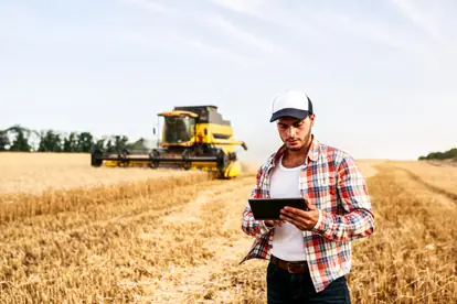 Landwirt nutzt ein Tablet zur Überwachung einer Ernte mit einem modernen Mähdrescher im Hintergrund.