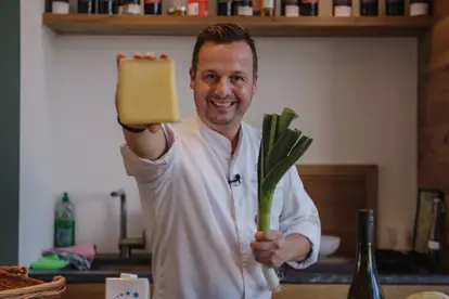 Ein Koch hält ein Stück Käse und einen Lauch in der Hand, bereit für die Koch-Challenge.
