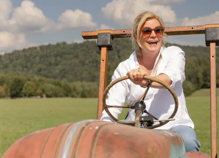 Eine lächelnde Frau fährt einen alten Traktor auf einem grünen Feld unter einem blauen Himmel.