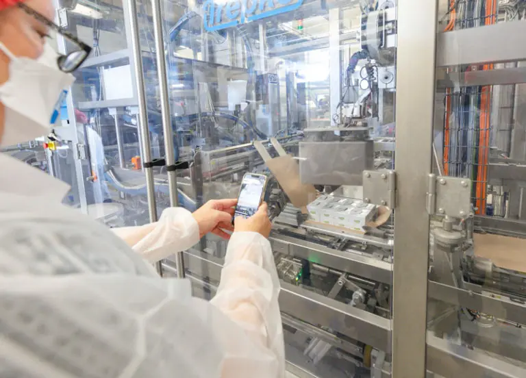 Eine Person in Schutzkleidung bedient ein Smartphone und überwacht eine automatisierte Verpackungsmaschine in einer Fabrik.