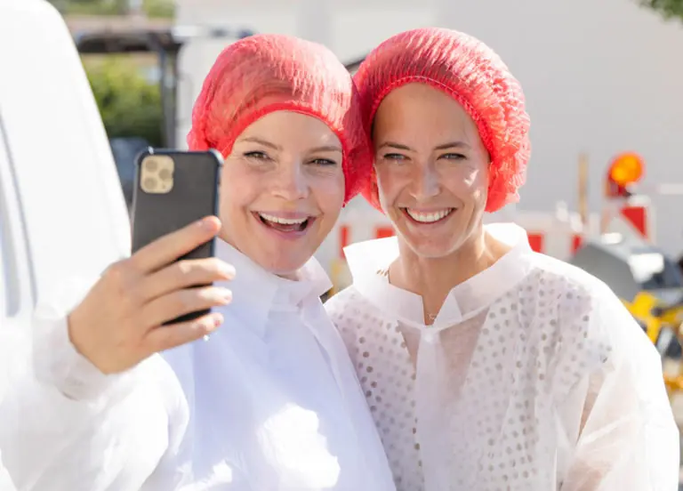Zwei lächelnde Frauen in Schutzkleidung und Haarnetzen machen ein Selfie im Freien.