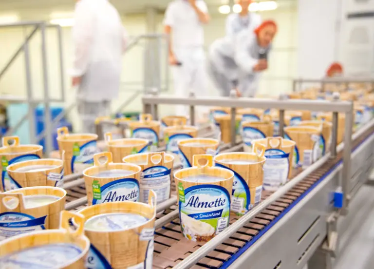 Produktionslinie mit Almette-Quarkbechern in einer Fabrik, im Hintergrund Mitarbeiter in Schutzkleidung.
