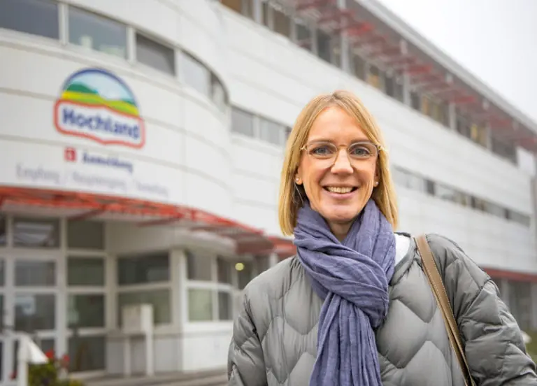 Eine lächelnde Frau mit Brille steht vor dem Hochland-Gebäude, das den Empfangsbereich zeigt.