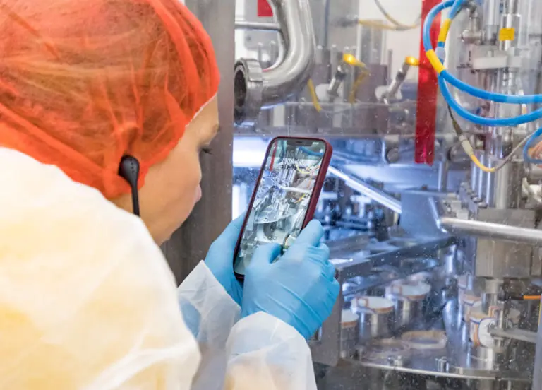 Eine Person in Schutzkleidung überwacht eine Produktionsmaschine mit einem Smartphone in einer industriellen Umgebung.