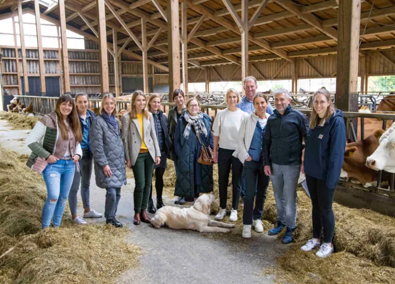 Gruppe von Menschen und einem Hund in einem Stall mit Kühen, umgeben von Heu und Holzstrukturen.