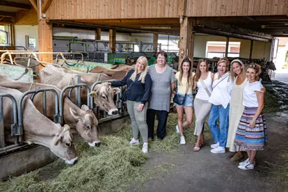 Sechs Frauen stehen in einem Stall neben Kühen und Heu, umgeben von ländlicher Atmosphäre und landwirtschaftlichem Ambiente.