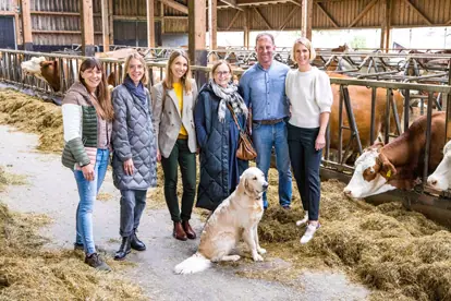 Sechs Personen und ein Hund stehen in einem Stall zwischen Kühen und Heu, lächelnd und in entspannter Atmosphäre.