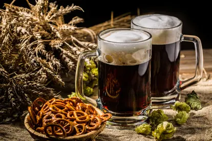 Zwei Gläser Hopfen Bier mit Brezeln und Hopfen neben einer Getreidebündel-Dekoration auf einem Holztisch.
