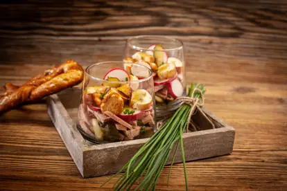 Knödelcarpaccio serviert in Gläsern mit Radieschen, Wurst und Kräutern, begleitet von einer Brezel auf Holztablett.