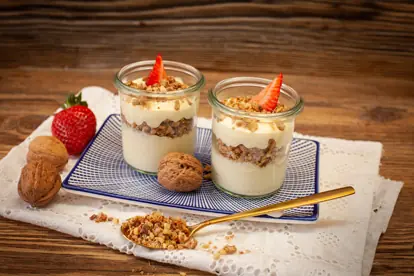 Zwei Gläser mit Knödelcarpaccio, Joghurt, Müsli und Erdbeeren auf einem dekorativen Teller mit Löffel und Nüssen.