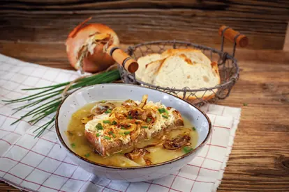 Knödelcarpaccio in einer Schüssel mit Brühe, Röstzwiebeln und frischem Brot, umgeben von Schnittlauch und Holzoberfläche.