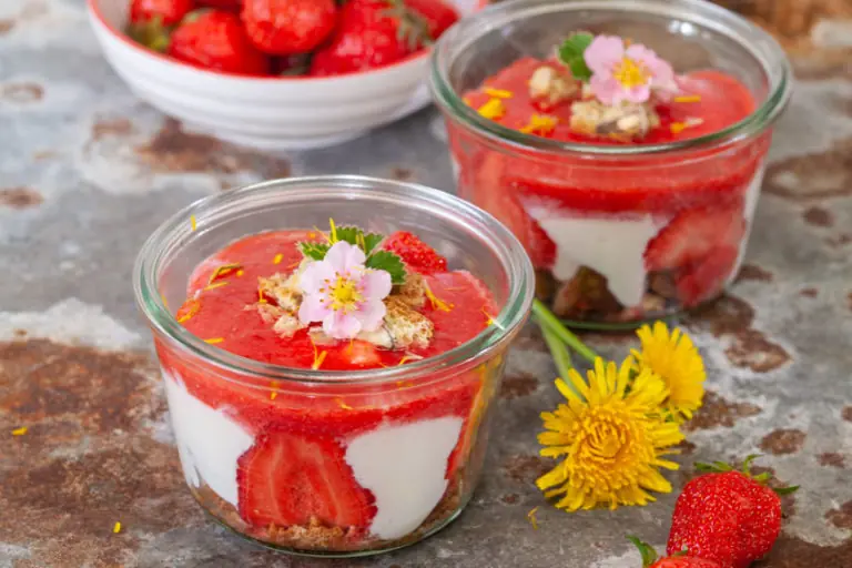 Erdbeerbecher mit frischen Erdbeeren, Joghurt und knusprigem Boden, dekoriert mit Blumen und Kräutern.