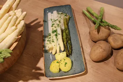 Weißer und grüner Spargel mit Buttersoße und Kartoffeln auf einem Teller, umgeben von frischem Spargel und Kartoffeln.