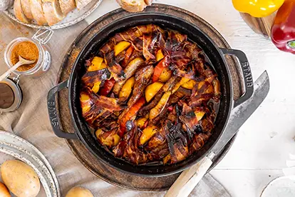 Schichtfleisch im Dutch Oven mit buntem Gemüse und knusprigem Bacon, serviert auf rustikalem Tisch.