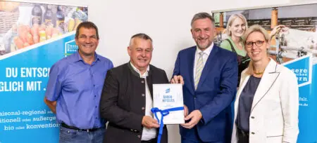 Vier Personen halten eine Urkunde in den Händen, umgeben von Plakaten mit regionalen Lebensmitteln und Landwirtschaft.