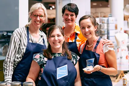 Vier Frauen in Schürzen lächeln fröhlich bei einer Veranstaltung zur Zubereitung von Dinkeldesserts mit Früchten.