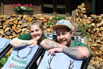 Zwei lächelnde Personen in Liegestühlen vor einem Holzstapel, die die Atmosphäre des "Kenzen"-Toasts genießen.
