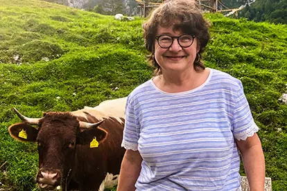 Eine lächelnde Frau steht auf einer Wiese neben einer Kuh, umgeben von grünen Bergen.
