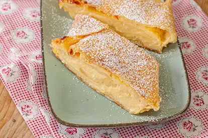 Zwei Stücke Topfenstrudel auf einem grünen Teller, bestäubt mit Puderzucker, serviert auf einem gemusterten Tuch.