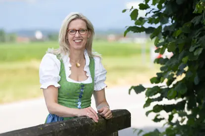 Eine Frau in traditioneller Tracht lächelt an einem Zaun, umgeben von einer ländlichen Kulisse, passend zum Verein.