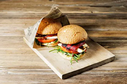 Zwei saftige Burger mit Grillkäse, Tomaten und Antipasti auf einem Holzbrett serviert.