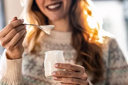 Eine lächelnde Frau hält einen Joghurtbecher und einen Löffel, was die Frage nach der Gesundheit von Milch verdeutlicht.
