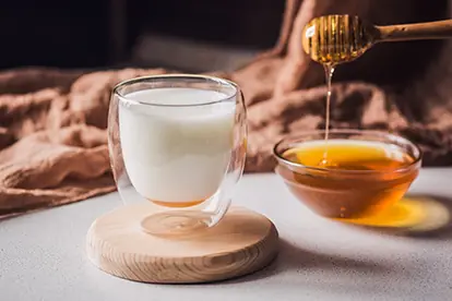 Ein Glas Milch auf einem Holzuntersetzer mit Honig in einer Schüssel, symbolisiert die Frage: Ist Milch gesund?