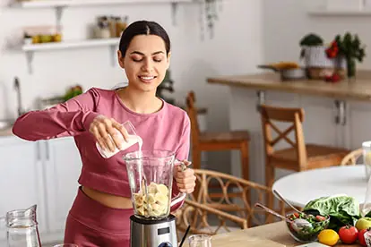 Eine Frau fügt Milch zu einem Mixer mit Bananen hinzu, um einen gesunden Smoothie zuzubereiten, passend zum Thema Milch gesund.