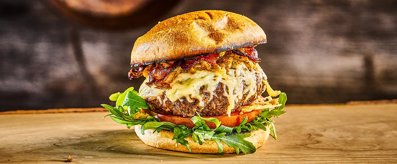 Saftiger Bayerischer Burger mit Käse, Speck, frischem Gemüse und einem knusprigen Brötchen auf einer Holzplatte.