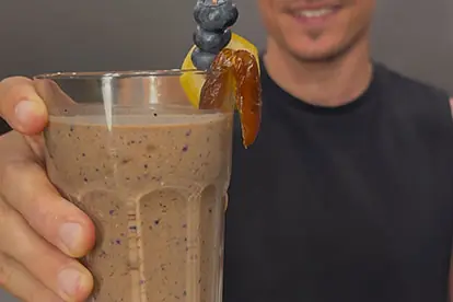 Ein Mann hält ein Glas Schoko-Bananen-Shake mit einer Garnitur aus Heidelbeeren und einer Orangenscheibe.