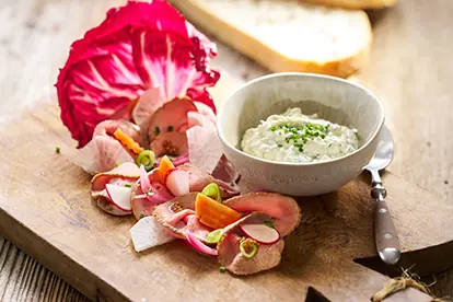 Kalbsbraten mit Kräuterquark, serviert mit buntem Gemüse und Brot auf einem Holzbrett.