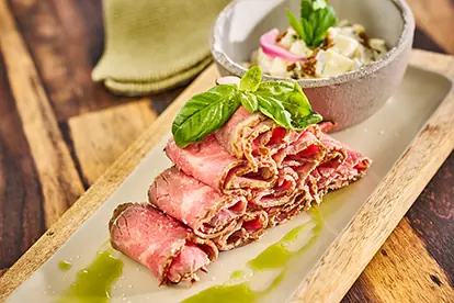 Saftiges Roastbeef auf einem Holzbrett, serviert mit einer Kräutersauce und einer Schüssel mit Salat.